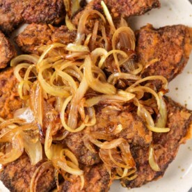 A plate of pan-fried beef heart and liver pieces topped with caramelized onions. The dark brown meats boast a crispy texture, while the golden onions glisten invitingly. This savory dish is elegantly presented on a light-colored plate.