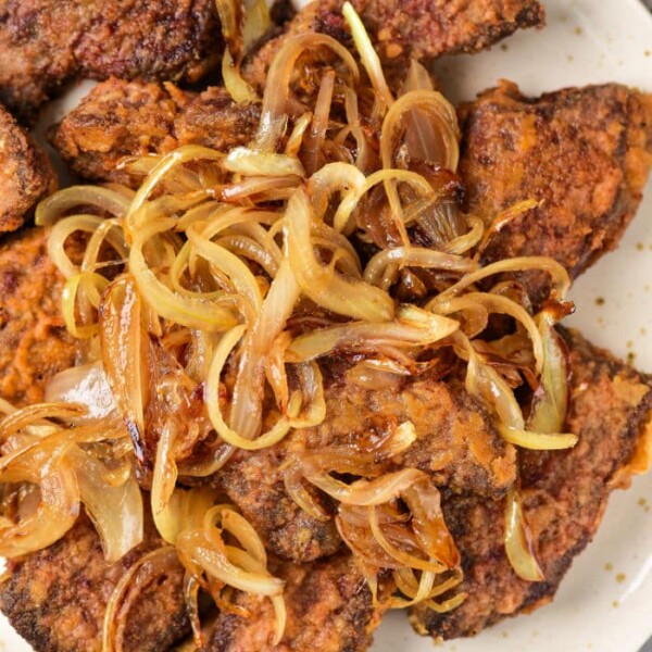 A plate of pan-fried beef heart and liver pieces topped with caramelized onions. The dark brown meats boast a crispy texture, while the golden onions glisten invitingly. This savory dish is elegantly presented on a light-colored plate.