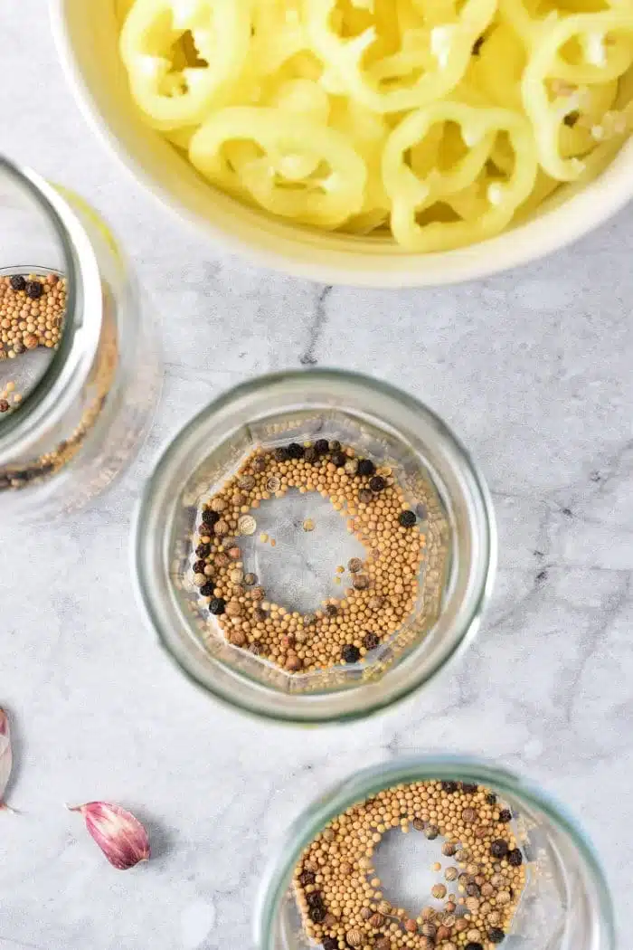 Three empty glass jars with mustard seeds and peppercorns sit on a marble surface, ready to preserve pickled fruits and vegetables. A bowl of sliced yellow peppers is nearby, while a clove of garlic peeks from the corner, setting the stage for culinary creativity.