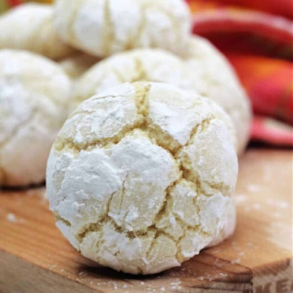 Close-up of a powdered lemon crinkle cookie on a wooden board, showcasing the delightful results of a cherished lemon crinkle cookie recipe. More cookies and a colorful cloth add vibrant charm to the background.