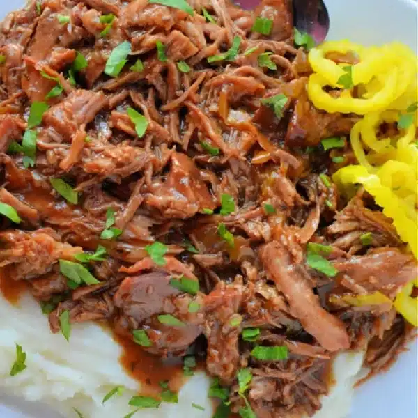 A plate of shredded pork, reminiscent of a Mississippi Pork Roast, served over mashed potatoes and garnished with chopped parsley. To the side, there's a serving of yellow pickled peppers. A spoon rests on the plate.