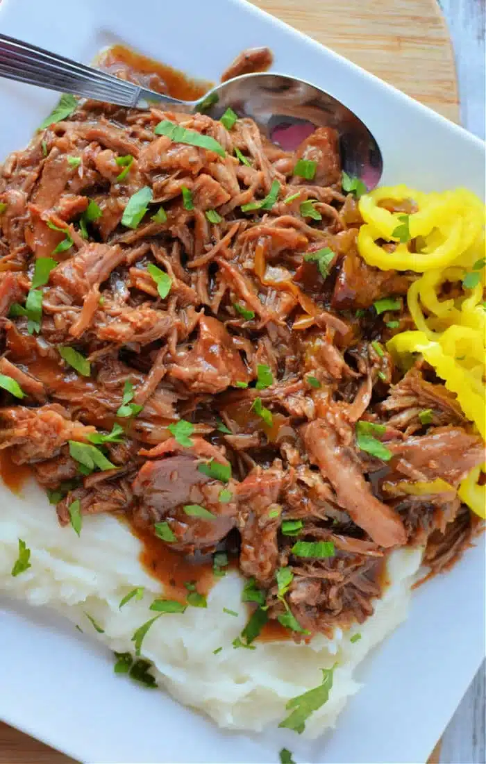 A plate of shredded pork, reminiscent of a Mississippi Pork Roast, served over mashed potatoes and garnished with chopped parsley. To the side, there's a serving of yellow pickled peppers. A spoon rests on the plate.