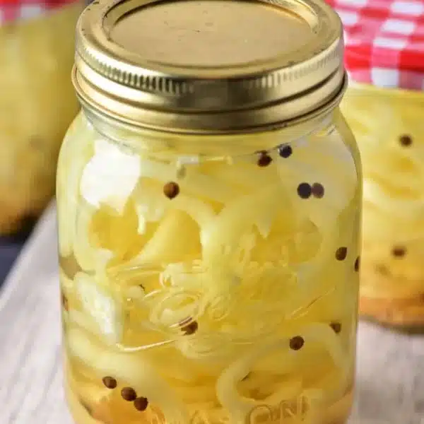 A mason jar filled with pickled banana pepper slices and mustard seeds, topped with a gold-colored lid, showcases the vibrant world of pickled fruits and vegetables. In the background, other jars, one adorned with a red and white checkered lid, are nestled on a wooden surface.