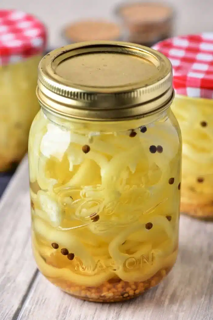 A mason jar filled with pickled banana pepper slices and mustard seeds, topped with a gold-colored lid, showcases the vibrant world of pickled fruits and vegetables. In the background, other jars, one adorned with a red and white checkered lid, are nestled on a wooden surface.