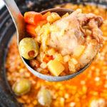 A ladle holding guisado de pollo with chicken, carrots, olives, and potatoes hovers above a simmering pot.