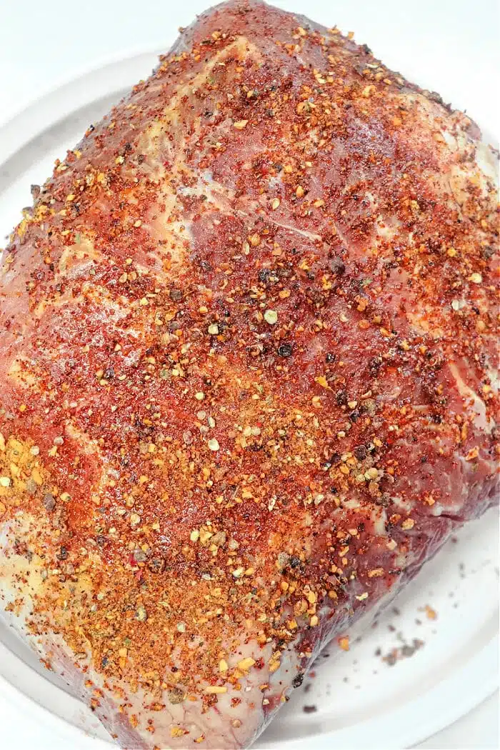 A raw smoked pork roast sits on a white plate, enveloped in a generous blend of red pepper flakes, ground pepper, and other fragrant herbs. The meat eagerly awaits its turn to be cooked or marinated to perfection.