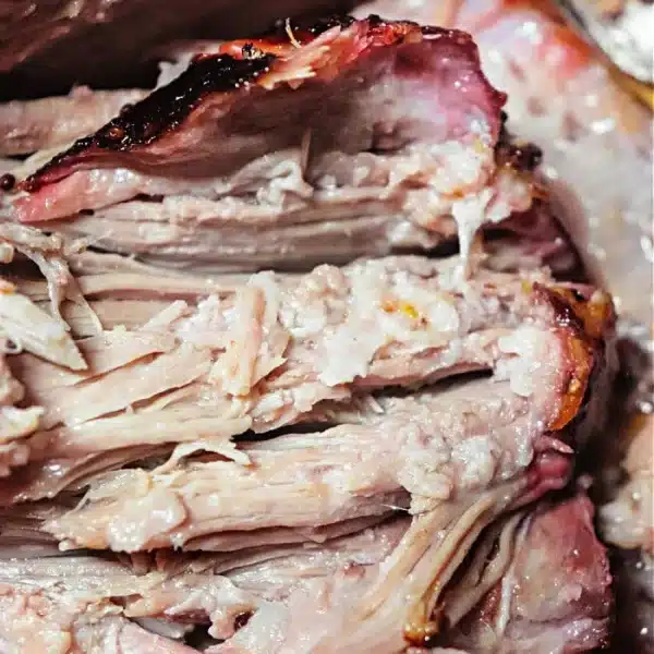 Close-up of sliced smoked pork roast showcasing tender, juicy meat and a reddish-brown crust. The texture appears soft and well-cooked, with visible layers and some charred edges. A fork is partially visible in the background.