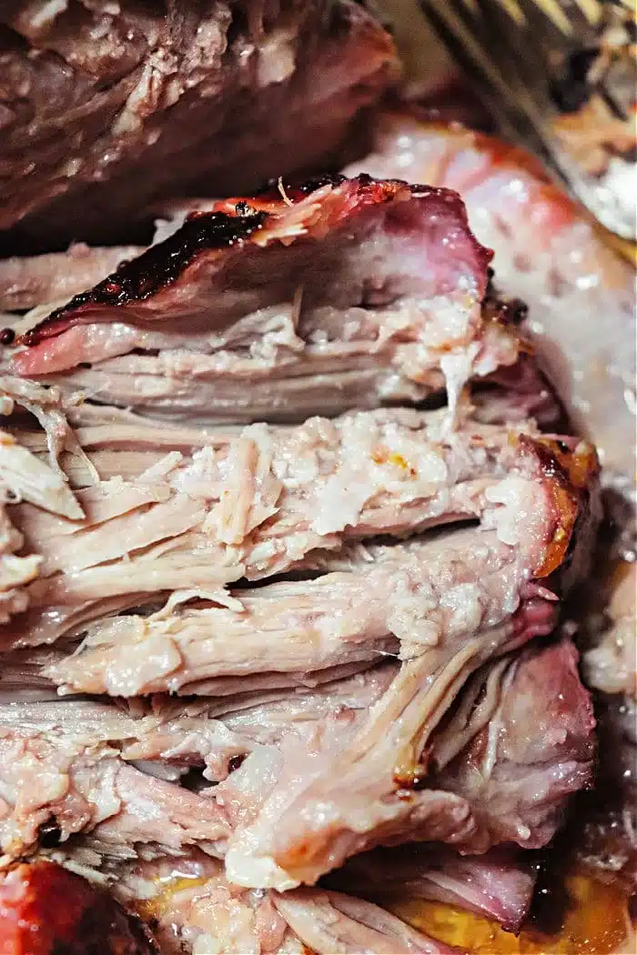 Close-up of sliced smoked pork roast showcasing tender, juicy meat and a reddish-brown crust. The texture appears soft and well-cooked, with visible layers and some charred edges. A fork is partially visible in the background.