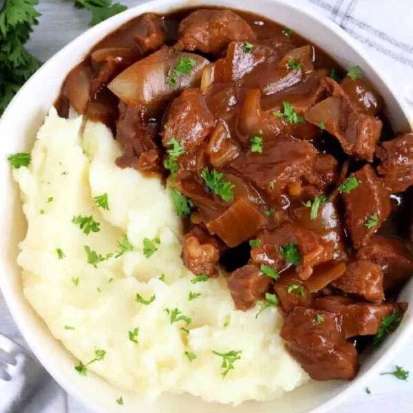 A savory bowl of mashed potatoes topped with beef stew is delicately garnished with parsley, sitting beside a neatly placed fork and napkin. It's a comforting dish that highlights the best in recipes using beef stew meat.