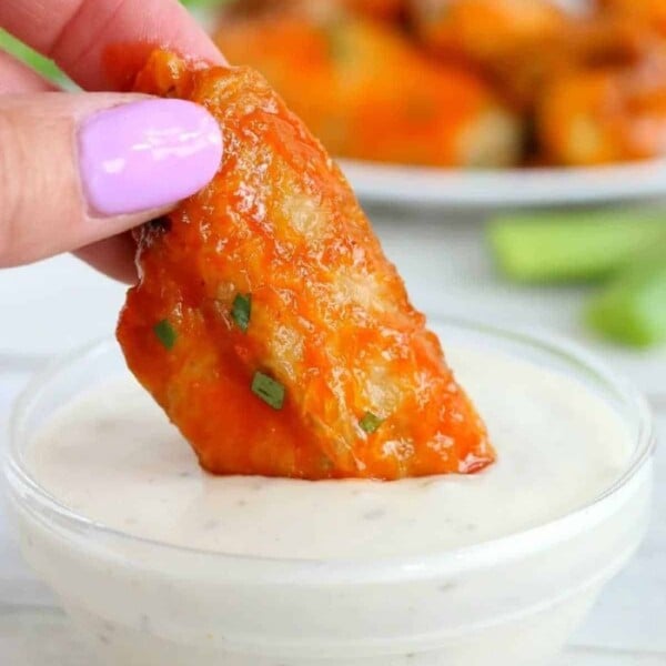 A hand dips a crispy buffalo chicken wing&mdash;made from frozen chicken wings in air fryer&mdash;into a bowl of ranch dressing, with more wings in the background.