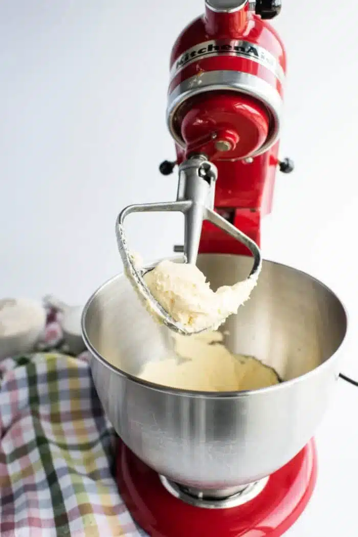 A red stand mixer with a metal bowl is in use, whipping a creamy mixture for homemade shortbread cookies with a paddle attachment. A plaid cloth is partially visible on the left side, and the background is a plain white wall.