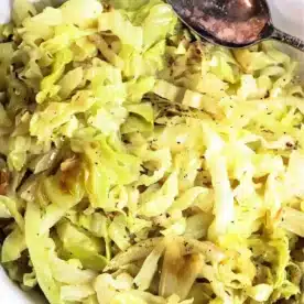 A white plate filled with slow cooker cabbage, seasoned with black pepper. A silver spoon rests on the edge of the plate.