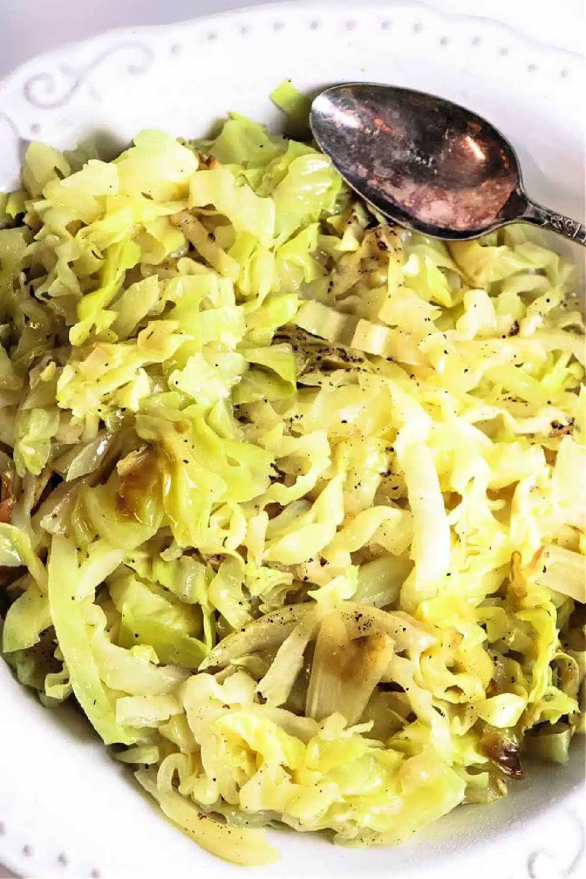 A white plate filled with slow cooker cabbage, seasoned with black pepper. A silver spoon rests on the edge of the plate.