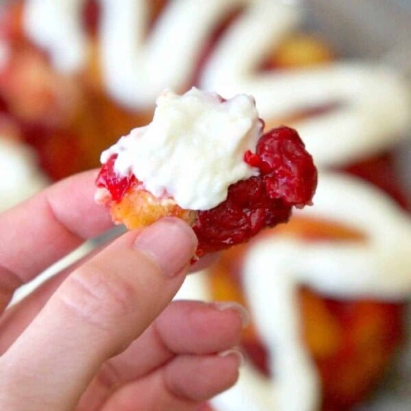 A hand holding a delightful piece of cherry cheesecake monkey bread, the pastry topped with whipped cream and vibrant red jam.