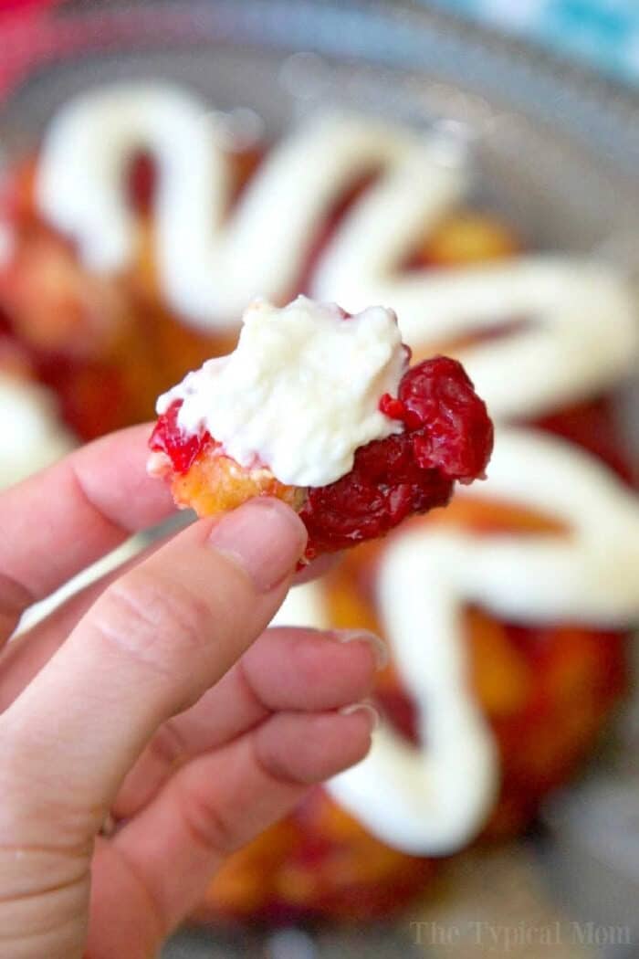 A hand holding a delightful piece of cherry cheesecake monkey bread, the pastry topped with whipped cream and vibrant red jam.