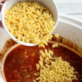 Pouring uncooked elbow macaroni into a simmering pot of crockpot chili mac with a rich tomato base adds hearty texture and delicious flavor.