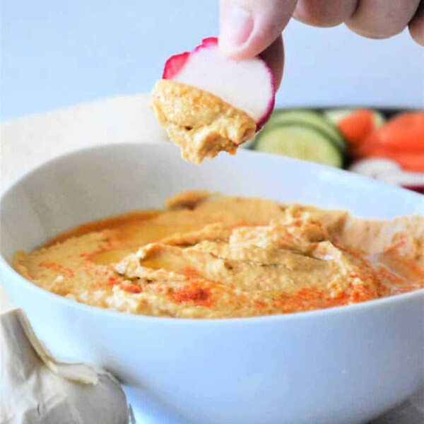 Hand dipping sliced radish into a creamy, easy hummus made with canned chickpeas, with garlic and fresh vegetables in the background.