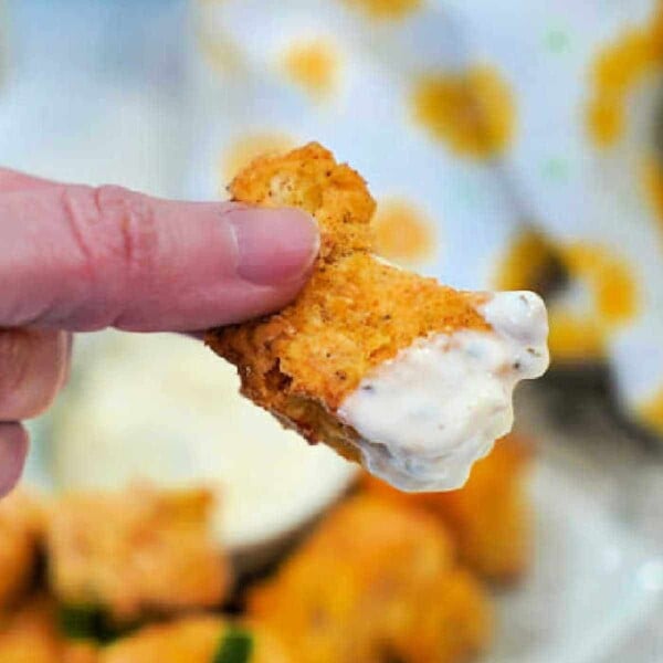 Close-up of a hand holding a breaded chicken nugget dipped in ranch sauce, with more nuggets and an air fryer hinting at perfectly crispy bites in the background.