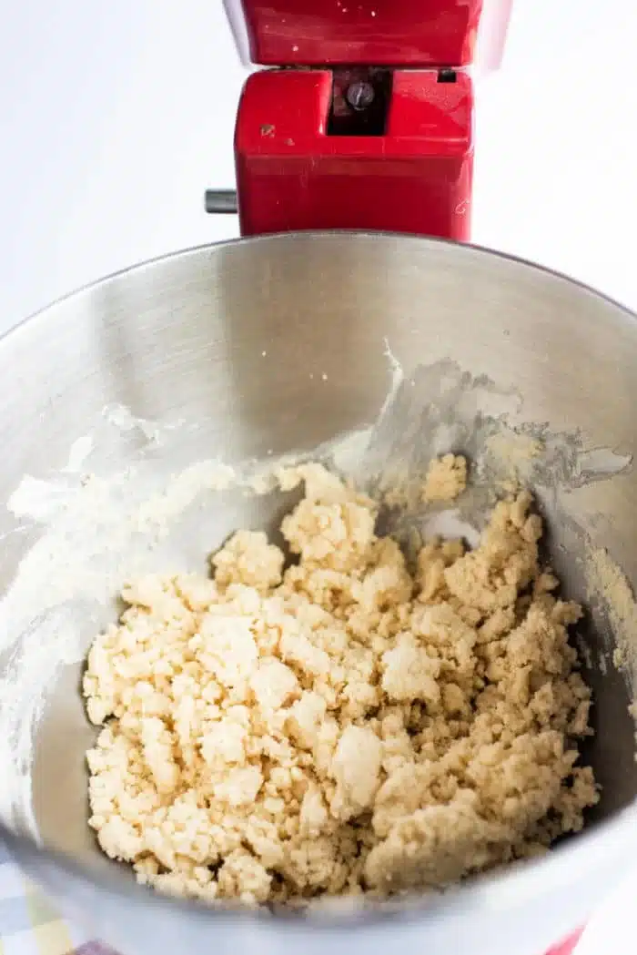 A close-up view of a mixing bowl attached to a red stand mixer, containing crumbly dough for homemade shortbread cookies being mixed. The dough appears light in color with a rough texture, hinting at its buttery goodness. The background is mostly white.