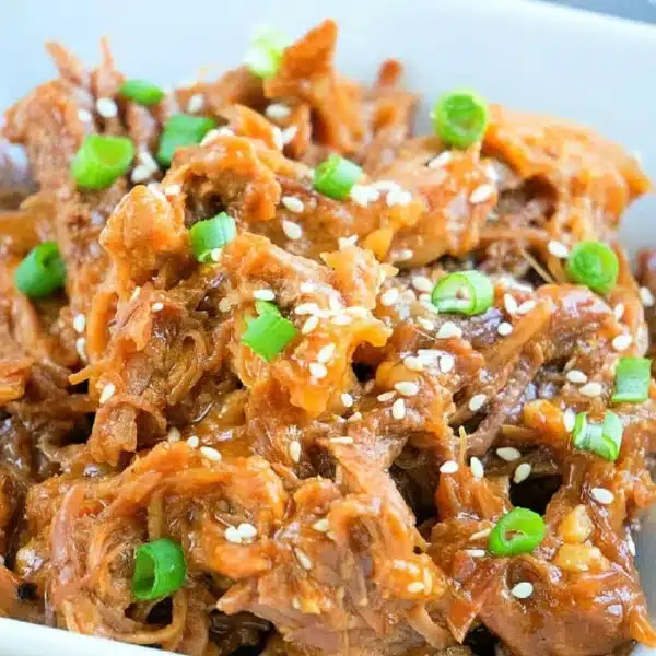 A white rectangular dish filled with Asian Instant Pot pulled pork, garnished with sliced green onions and sesame seeds. The shredded meat appears to be coated in a savory sauce.