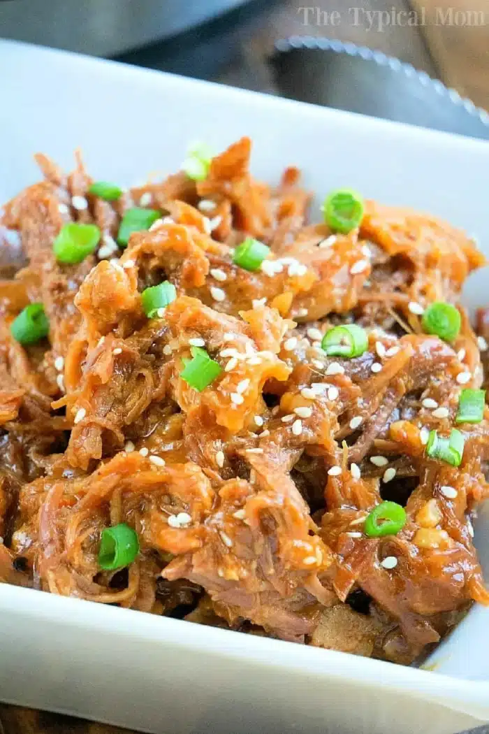 A white rectangular dish filled with Asian Instant Pot pulled pork, garnished with sliced green onions and sesame seeds. The shredded meat appears to be coated in a savory sauce.