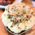 Whole pressure cooker cauliflower topped with herbs and cheese sits on a rack, with grilled meat in the background.