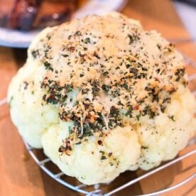 Whole pressure cooker cauliflower topped with herbs and cheese sits on a rack, with grilled meat in the background.