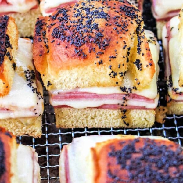 Close-up of baked slider sandwiches with ham and melted cheese, topped with poppy seeds, on a wire rack. The golden-brown bread has a slightly crispy texture, reminiscent of air fryer sandwiches.