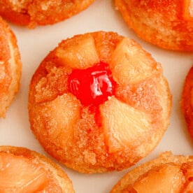 Close-up of individual pineapple upside-down cupcakes. Each cupcake showcases pineapple chunks arranged in a circular pattern, crowned with a bright red cherry at the center. The cupcakes boast a golden-brown caramelized surface.