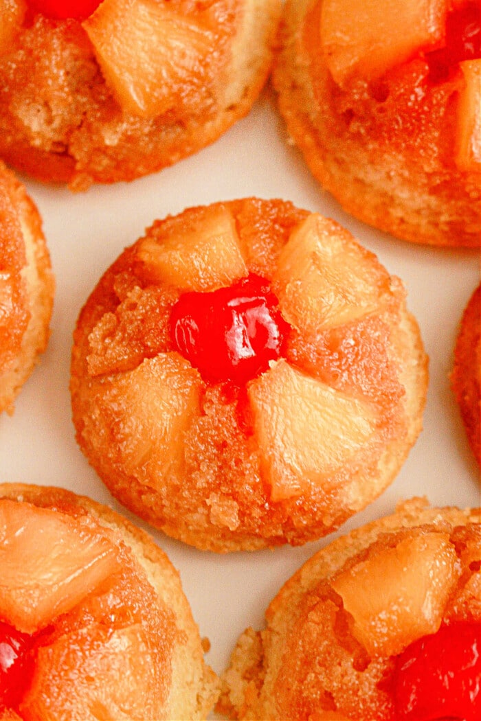 Close-up of individual pineapple upside-down cupcakes. Each cupcake showcases pineapple chunks arranged in a circular pattern, crowned with a bright red cherry at the center. The cupcakes boast a golden-brown caramelized surface.