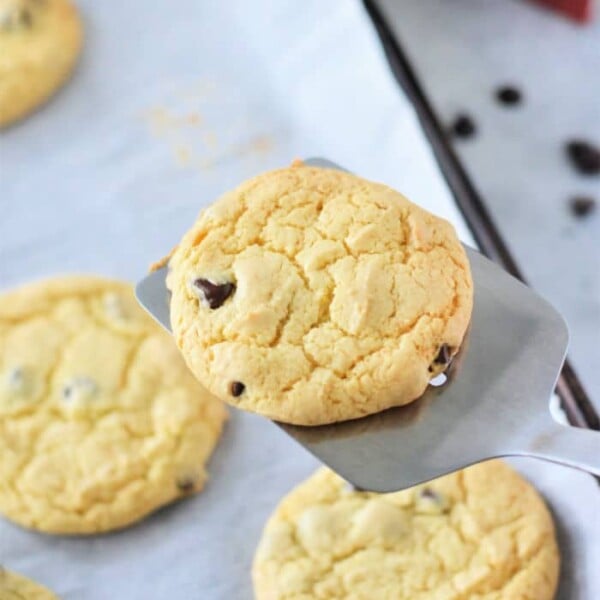 A spatula delicately lifts a chocolate chip cookie, revealing a tray of freshly baked delights on parchment paper, reminiscent of the sweetness you'd find in yellow cake mix cookies.