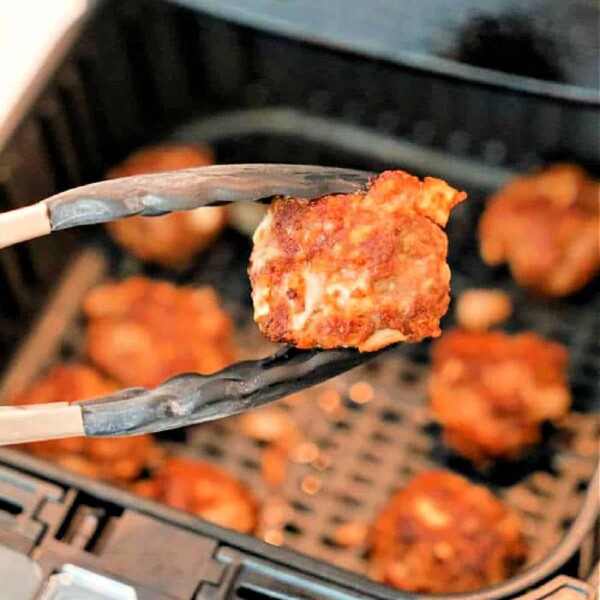 Close-up of tongs holding a piece of cooked chicken in front of an air fryer. Several other pieces, reminiscent of perfectly browned and crispy meatballs straight from the air fryer, are visible in the basket in the background.