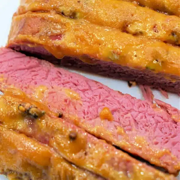 A close-up of sliced corned beef topped with mustard, displaying a pink, juicy interior. The texture appears tender, and the mustard sauce has a slightly golden, bubbly appearance. The slices are arranged neatly on a white surface, showcasing how to cook corned beef in the oven perfectly.