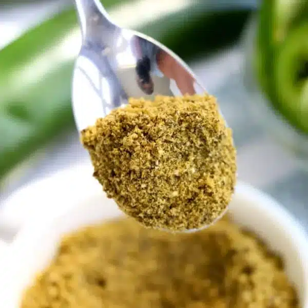 A close-up image of a spoonful of vibrant green jalapeno powder held above a white bowl filled with the same fiery seasoning. In the background, blurred green peppers are visible, hinting at the spice's origin and adding depth to this flavorful scene.