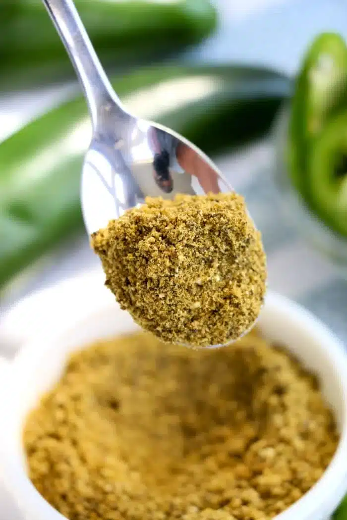 A close-up image of a spoonful of vibrant green jalapeno powder held above a white bowl filled with the same fiery seasoning. In the background, blurred green peppers are visible, hinting at the spice's origin and adding depth to this flavorful scene.