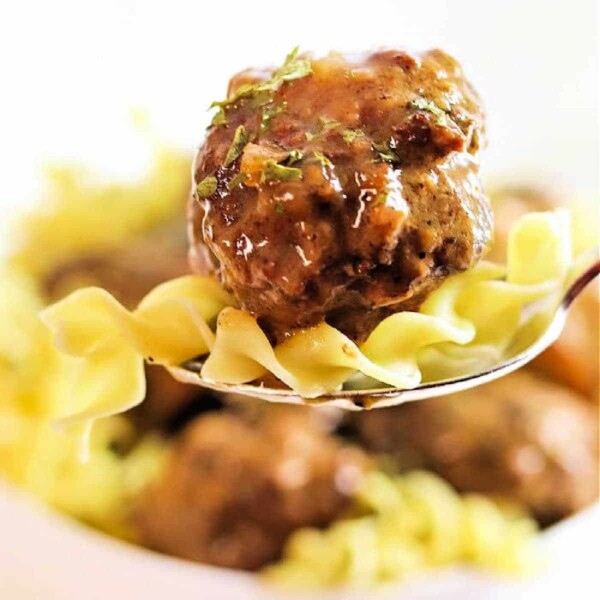 A fork with juicy crockpot meatballs and noodles, garnished with fresh herbs, stands out against a blurred background.