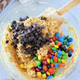 A clear glass bowl holds cookie dough mixed with chocolate chips and colorful candy-coated chocolates, ready to become delicious cookie bars from cake mix. A spatula rests in the bowl, while a chocolate cake mix box peeks out from the background on a light gray surface.