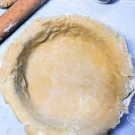 A rolled-out pot pie crust is placed in a round pie dish on a light blue surface. A wooden rolling pin and a small cup of flour are visible nearby, ready for adjustments. The dough has a slightly uneven edge, adding to its homemade charm.