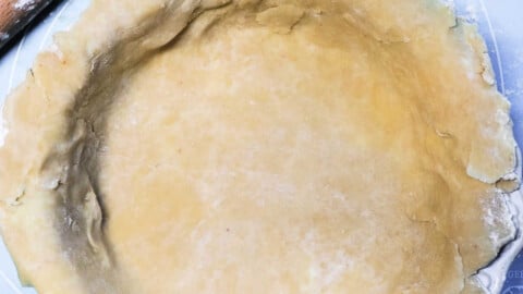 A rolled-out pot pie crust is placed in a round pie dish on a light blue surface. A wooden rolling pin and a small cup of flour are visible nearby, ready for adjustments. The dough has a slightly uneven edge, adding to its homemade charm.
