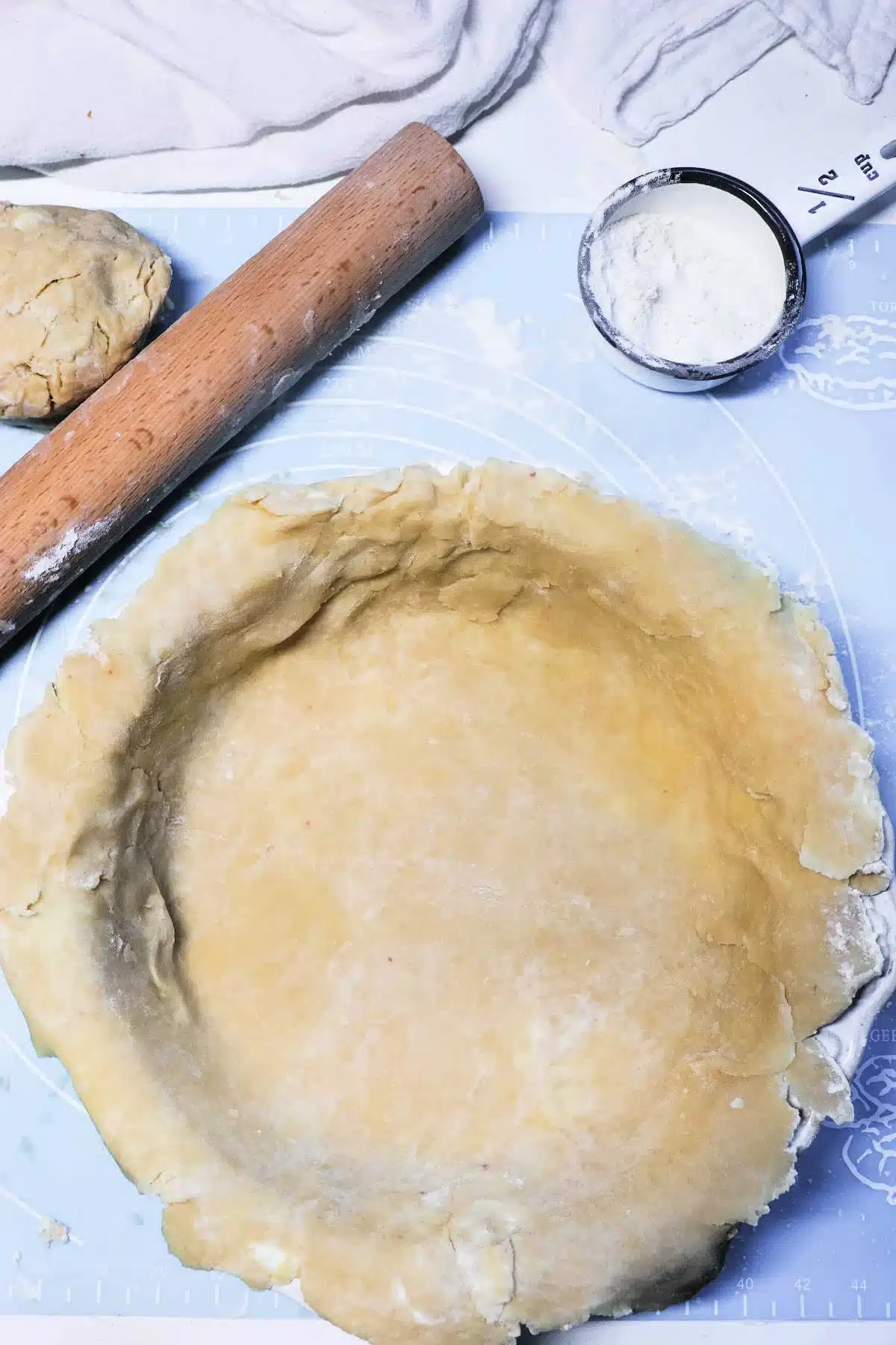 A rolled-out pot pie crust is placed in a pie dish on a blue work surface. Nearby are a wooden rolling pin, a ball of dough, and a tin measuring cup with flour. White fabric is in the background.