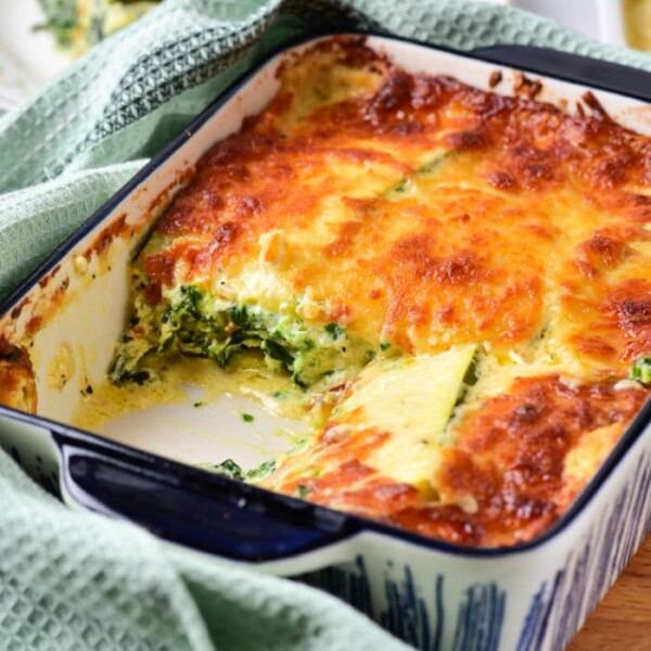 A baked vegetable lasagna with white sauce, sits invitingly in a dish with a piece missing, surrounded by a green towel.