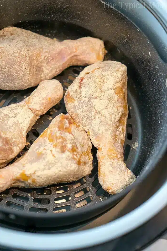 Close-up of four raw, breaded chicken drumsticks placed in the basket of an air fryer, ready to be cooked. The chicken is lightly coated with a flour mixture, perfect for an easy air fryer fried chicken recipe. The air fryer basket's black mesh is visible underneath the drumsticks.