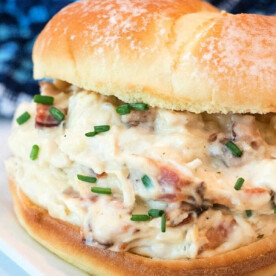 A creamy crockpot crack chicken bacon sandwich on a toasted bun, garnished with chopped chives, served on a white plate with a blue patterned cloth in the background.