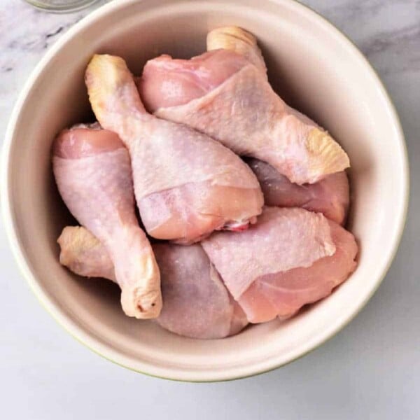 Raw chicken drumsticks rest in a white bowl on the marble countertop, awaiting transformation. Nearby, sliced garlic sits ready in a small dish. Perfect for those wondering how long to boil chicken legs for the best flavor and tenderness.