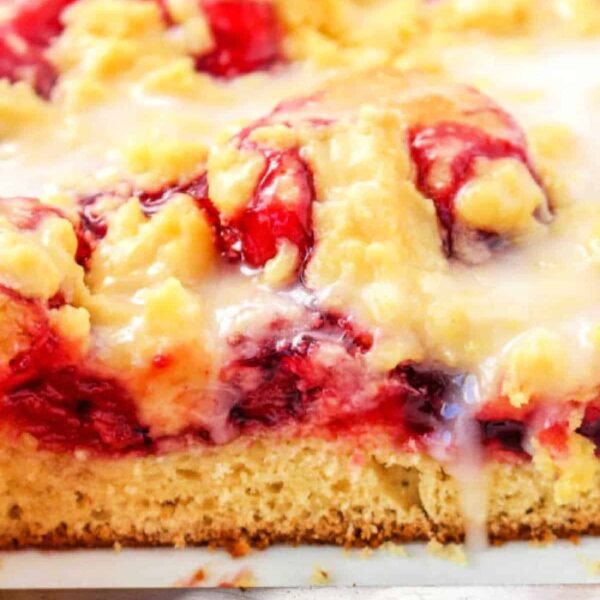 Close-up of a cherry coffee cake, studded with berries and drizzled with icing, resting on a metal tray.