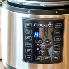 A sleek silver Crockpot with a digital display shows the letter "H." The front panel features buttons for various Express settings like meat/stew, beans/chili, yogurt, and slow cook. A spoon rests in the open lid, ready for your culinary creations.