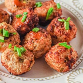 A white decorative plate features several golden brown air fryer chicken meatballs, garnished with chopped green onions. The perfectly cooked meatballs are arranged closely together for a delicious presentation.