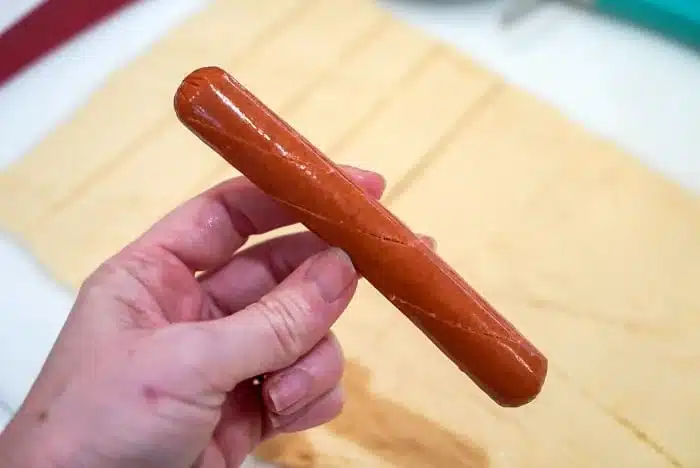 A hand holds a raw hot dog ready to wrap in crescent dough ready to wrap and bake.