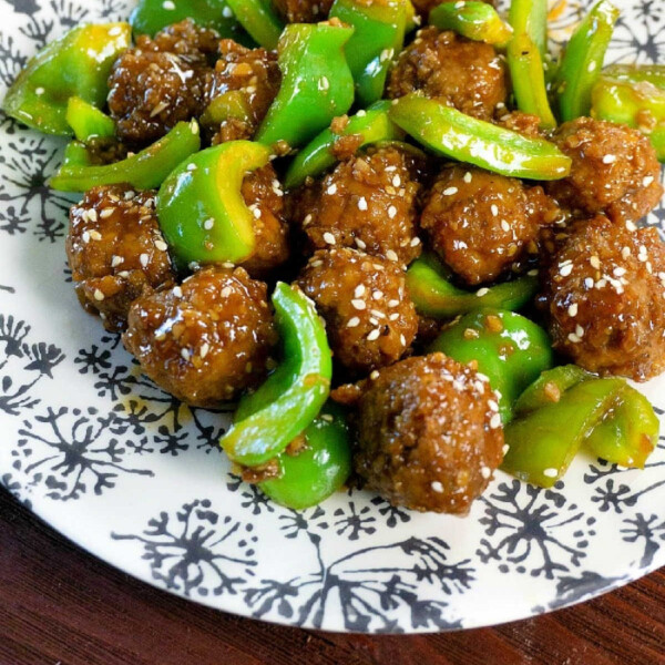 A plate with glazed instant pot meatballs mixed with sliced green bell peppers, sprinkled with sesame seeds, elegantly served on a white plate featuring a black floral pattern. The presentation rests on a rustic wooden surface.