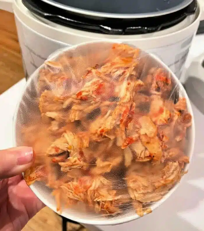 A person holds a plastic-covered bowl filled with seasoned, shredded chicken, perfect for leftover chicken recipes. An Instant Pot can be seen in the background, ready to create delightful dishes from these tender morsels.